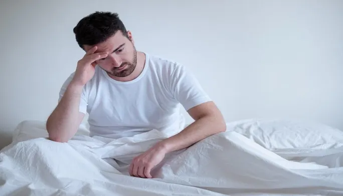 Man sitting on bed with a worried expression, reflecting emotional distress related to erectile dysfunction and seeking solutions through pelvic floor physical therapy.